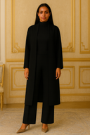 Woman in elegant black outfit standing in an ornate room with beige walls and a matching chair.