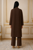 Model wearing brown long coat and trousers, standing against ornate wall, back view in elegant interior setting.