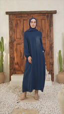 Model wearing blue sheath abaya maxi dress with kimono sleeves and pockets, standing on white stones. Elegant modest fashion.