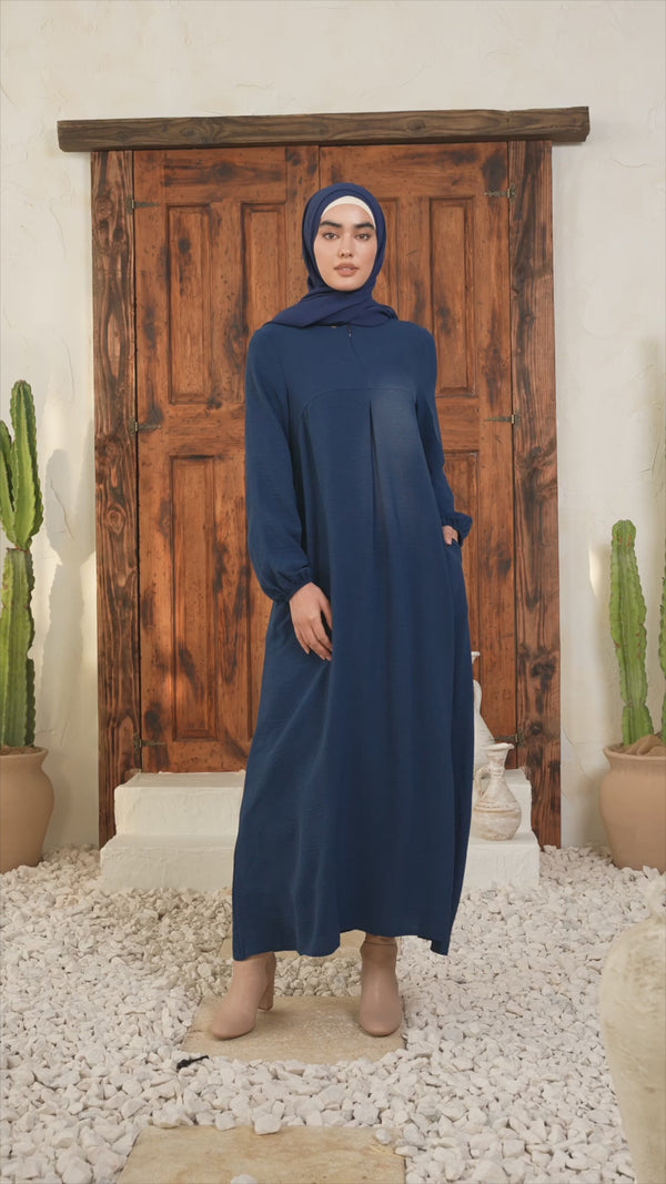 Model wearing blue sheath abaya maxi dress with kimono sleeves and pockets, standing on white stones. Elegant modest fashion.